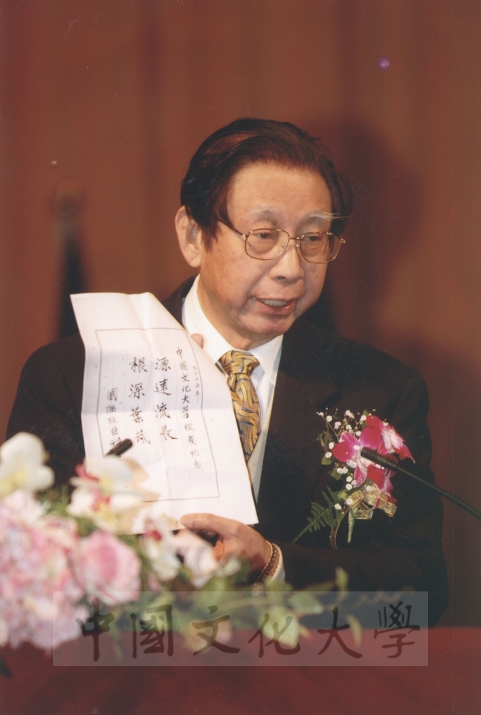 2005年3月1日建校43週年校慶典禮上董事長張鏡湖出示國際歐亞科學院祝賀本校校慶賀詞「源遠流長，根深葉茂」的圖檔，第1張，共1張